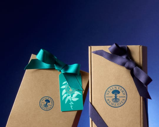 Two kraft paper gift boxes from Neal's Yard Apothecary, one with a teal ribbon and tag, and one with a dark purple ribbon, set against a dark blue gradient background.