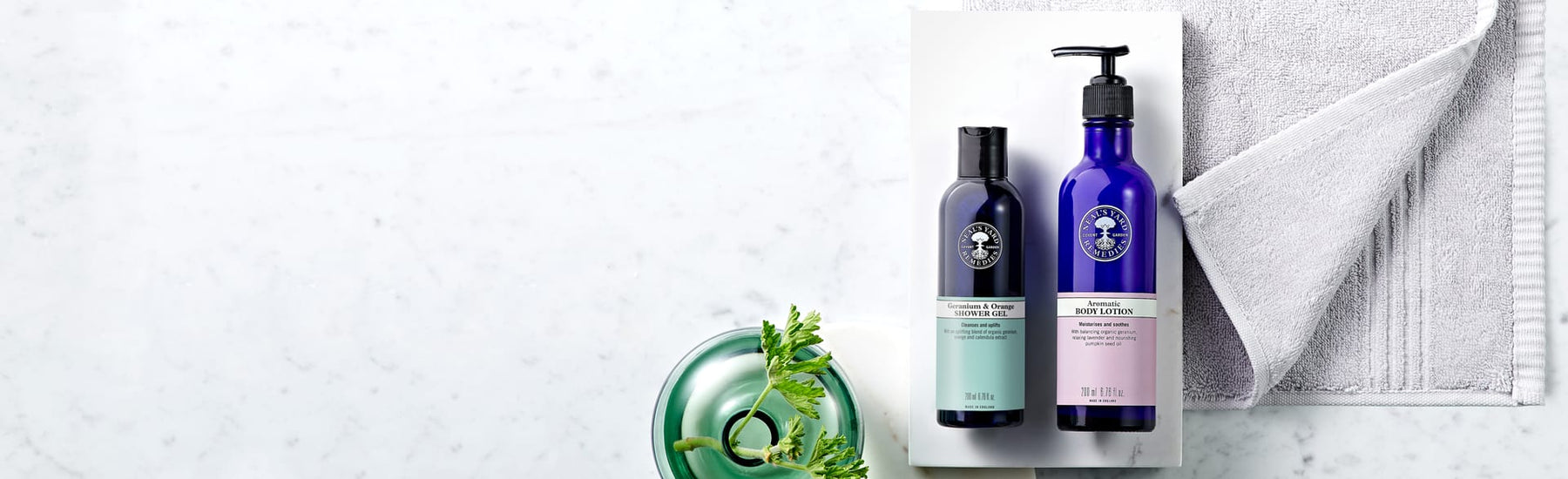 Picture of a shower gel and a body lotion a grey towel and plant in vase by Neal's Yard Remedies
