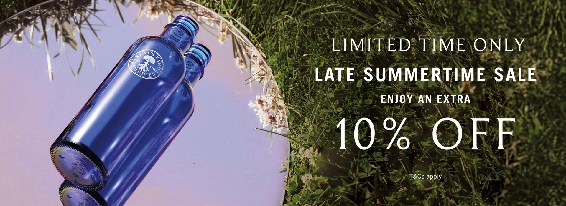 A blue glass bottle shown on a split background with a purple reflective section on the left and green grass on the right, alongside sale text reading 'LIMITED TIME ONLY LATE SUMMERTIME SALE ENJOY AN EXTRA 10% OFF'.
